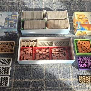 Puede incluir: Vista aérea de un juego de mesa, "Railroad Tiles", con varios componentes. El juego incluye cajas, cartas, fichas y bolsas con cordón en azul, amarillo y verde. El tema del juego parece ser una ciudad o un paisaje.