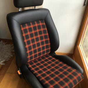 Może przedstawiać: Czarny fotel samochodowy z zagłówkiem. Siedzisko ma czerwono-żółto-czarny wzór w kratę. Siedzisko wykonane jest ze skóry i posiada klamrę pasa bezpieczeństwa.