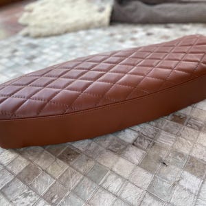 Op de afbeelding: Een bruin motorzadel met een ruitvormig stikselpatroon. De zitting is ovaal van vorm en gemaakt van een glad, leerachtig materiaal. De randen zijn afgerond en de zitting lijkt nieuw.