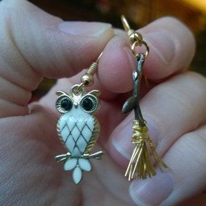 May include: A pair of gold earrings, one featuring a white owl with black eyes and the other a gold twig with a gold broom attached.