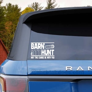 May include: A blue SUV with a white vinyl sticker on the back window that says "Barn Hunt May the Course Be With You".