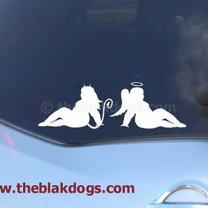 May include: White vinyl decal of two figures, one with angel wings and a halo, the other with a tail and horns, both in a reclining pose. The decal is on a blue car window. The text "www.theblakdogs.com" is at the bottom of the image.