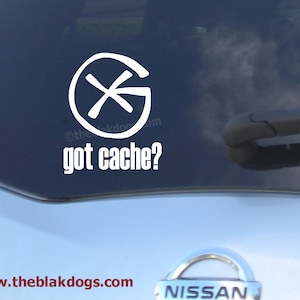 May include: White vinyl sticker on a car window with a compass symbol and the text "got cache?"  The sticker is on a silver car with a Nissan emblem.