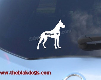 Puede incluir: Calcomanía de vinilo blanco con la silueta de un perro Gran Danés con el nombre "Angus" y un corazón dentro de la silueta. La calcomanía está en la ventana trasera de un coche.