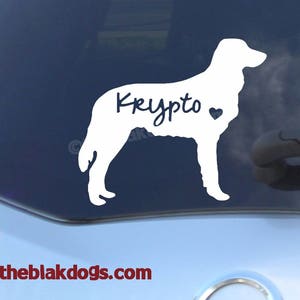 May include: White vinyl decal of a dog silhouette with the name "Krypto" in cursive and a heart symbol. The decal is displayed on a car window. The website address www.theblakdogs.com is visible at the bottom.