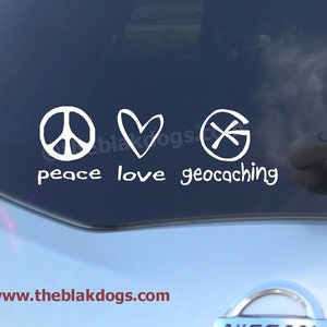 May include: White peace sign, heart, and compass symbol with the text "peace love geocaching" on a blue car window.
