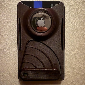May include: A brown cardholder with an Apple AirTag in the center. The cardholder has a textured surface with curved lines and a small textured grip at the bottom. A credit card is visible at the top.