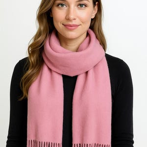 May include: A pale pink scarf with a fringed edge. The scarf is draped around the neck and shoulders. The person is wearing a black long-sleeved top.
