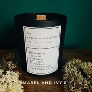 May include: A black candle with a wooden lid and a white label. The label reads "Orange Blossom, Neroli & Jasmine" and "burn time - 25 hours." The candle is 300g soy wax. The text "60 Years =" is also on the label.