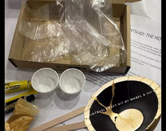 May include: Kintsugi craft kit featuring a black and gold ceramic bowl, two small white cups, two tubes of resin, a small bag of gold powder, and two wooden sticks. The kit includes instructions for repairing broken ceramics with gold.
