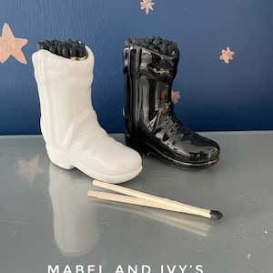 May include: A pair of ceramic cowboy boot match holders, one white and one black, filled with matches. The boots are sitting on a gray surface with two matches laying in front of them. The text "MABEL AND IVY'S" is printed on the surface.