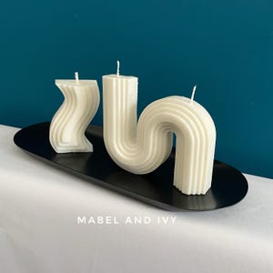 May include: Three white, geometrically shaped candles on a black oval tray. The candles are in the shape of a wave, a curved line, and a curved line with a loop. The tray has the text "Mabel and Ivy" printed on it.