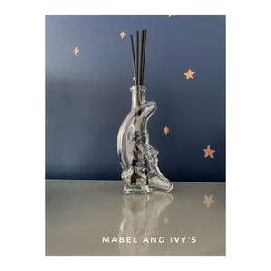 Könnte beinhalten: Eine klare Glasdiffuserflasche in Form eines Halbmondes mit schwarzen Diffusorsticks. Die Flasche steht auf einer weißen Oberfläche mit blauem Hintergrund.