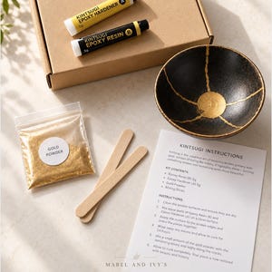 Puede incluir: Un kit de reparación Kintsugi que contiene una caja de cartón marrón, dos tubos de 5g etiquetados como Kintsugi Epoxy Hardener A y Epoxy Resin B, una bolsa de polvo dorado, dos palitos de madera, una hoja de instrucciones y un cuenco negro.
