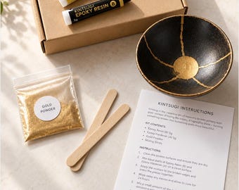 May include: A Kintsugi repair kit containing a brown cardboard box, two 5g tubes labelled Kintsugi Epoxy Hardener A and Epoxy Resin B, a bag of gold powder, two wooden mixing sticks, an instruction sheet, and a black bowl with gold cracks.
