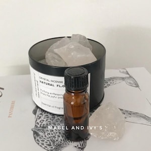 Può includere: Un contenitore nero con cristalli bianchi all'interno, etichettato "Crystal Incense Natural FL.O. Purifying and Relaxing Enjoy the joyful process Essential oil fragrance". Una piccola bottiglia di vetro marrone con un tappo nero è accanto al contenitore. Il contenitore e la bottiglia sono su una superficie bianca con una stampa di animali in bianco e nero.