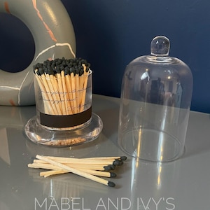 May include: A glass jar filled with black-tipped matches under a clear glass cloche. The jar has a black label with the text "You are playing with matches I have a paper heart By Mabel".