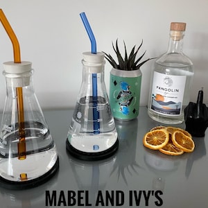Könnte beinhalten: Zwei Glasbecher mit orangefarbenen und blauen Strohhalmen, eine durchsichtige Glasflasche mit dem Etikett "Pangolin", eine Dose mit dem Text "India Pale Ale" und getrocknete Orangenscheiben auf einer grauen Oberfläche. Der Text "Mabel and Ivy's" befindet sich am unteren Rand des Bildes.
