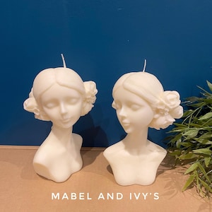 Könnte beinhalten: Zwei weiße, skulpturale Büstenkerzen mit floralen Details. Die Kerzen stehen auf einer braunen Oberfläche mit grünem Laub im Hintergrund. Der Text "MABEL AND IVY'S" ist auf der Oberfläche aufgedruckt.