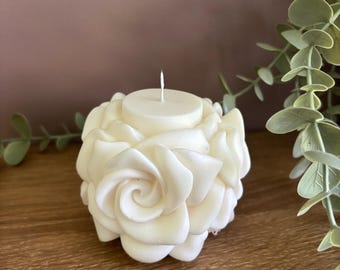 Può includere: Candela a forma di rosa color crema con uno stoppino centrale. La candela presenta petali dettagliati, creando un design floreale realistico. La base della candela è visibile e il testo "MABEL AND IVY'S" è stampato sulla superficie di legno.