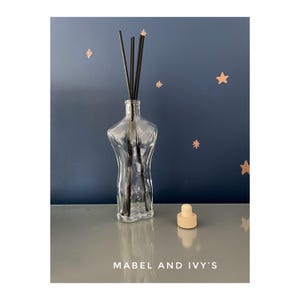 Pode incluir: Uma garrafa de difusor de vidro transparente com uma superfície texturizada e uma rolha de madeira. Varetas difusoras pretas estão na garrafa. A garrafa está sobre uma superfície cinza com o texto "MABEL AND IVY'S" abaixo.