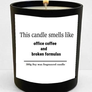 May include: A black candle with a white label. The label reads "This candle smells like office coffee and broken formulas." The candle is lit, and the label states it is a 300g soy wax fragranced candle.