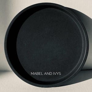 Puede incluir: Una bandeja redonda negra con un borde elevado. La bandeja tiene el texto "MABEL AND IVYS" impreso en la parte inferior.