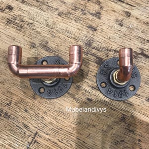 Puede incluir: Dos perchas industriales de tubería de cobre montadas sobre placas de metal negro. Las placas tienen un diseño circular con el número "1914" y las letras "DS" estampadas. Las perchas tienen forma de U.