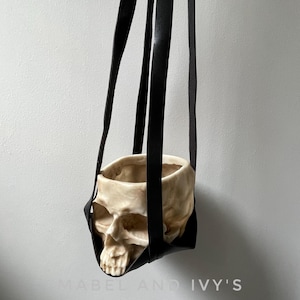 Puede incluir: Un macetero colgante de cuero negro con forma de calavera. El macetero está vacío y listo para ser llenado con plantas.