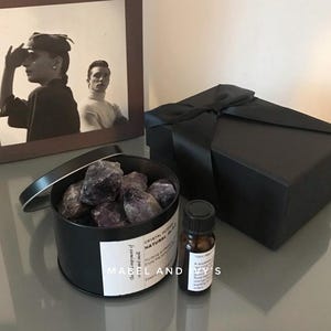 Op de afbeelding: Een zwarte doos met een satijnen lint eromheen gebonden. De doos staat naast een zwarte blik met amethistkristallen en een klein flesje etherische olie. Het etiket op de blik staat "Crystal incense natural plus" en het etiket op de fles staat "Mabel and Ivy's".