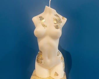 Puede incluir: Una vela blanca con forma de torso con detalles de pan de oro. La vela está sobre un soporte dorado, con el texto "MABEL AND IVY'S" en la base. El fondo es azul liso.