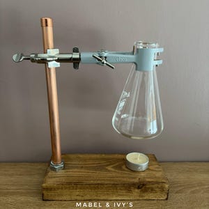 May include: A copper pipe and wooden base with a glass beaker held by a clamp. A small candle sits on the base. The base is labelled "MABEL & IVY'S".