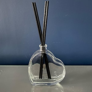 Könnte beinhalten: Eine klare Glasflasche in Herzform mit schwarzen Diffusorstäbchen. Die Flasche steht auf einer silbernen Oberfläche.