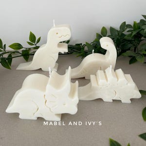 May include: Four white wax candles shaped like dinosaurs. The candles are on a light brown surface with green plants in the background. The candles are labeled "Mabel and Ivy's".