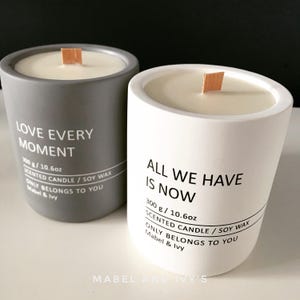 May include: Two grey and white scented candles with wooden wicks. The grey candle says "Love Every Moment" and the white candle says "All We Have Is Now". Both candles are 300g / 10.6oz and are made with soy wax. The candles are from Mabel & Ivy.