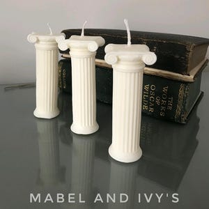 Puede incluir: Tres velas blancas en forma de columna con detalles dorados. Las velas están dispuestas sobre una mesa con una pila de libros en el fondo. Los libros tienen el título "The Works of Oscar Wilde" en la portada.