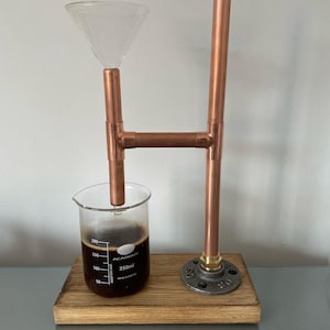 Puede incluir: Cafetera de tubería de cobre con un vaso de precipitados de vidrio y un embudo. El vaso de precipitados tiene marcas para 50, 100, 150 y 200 mililitros. La base está hecha de madera con un accesorio de tubería de metal.