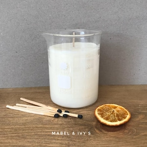 Puede incluir: Una vela blanca en un vaso de precipitados de vidrio transparente con una mecha blanca. El vaso de precipitados tiene marcas para medir en mililitros. Hay cerillas y una rodaja de naranja seca sobre una superficie de madera.