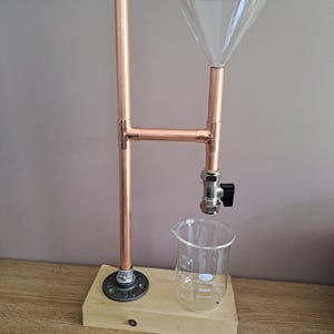 Puede incluir: Un soporte de tubería de cobre con un embudo de vidrio, un vaso de precipitados de vidrio y una válvula. El soporte está montado sobre una base de madera.