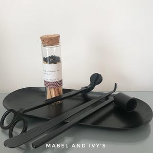 K&ouml;nnte beinhalten: Ein schwarzer Kerzenl&ouml;scher, ein schwarzer Kerzen-Docht-Trimmer und ein Glasgef&auml;&szlig; gef&uuml;llt mit schwarzen Streichh&ouml;lzern auf einem schwarzen Tablett. Das Tablett tr&auml;gt den Text "Mabel and Ivy's".