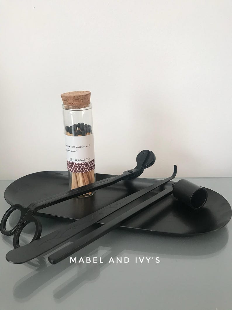 K&ouml;nnte beinhalten: Ein schwarzer Kerzenl&ouml;scher, ein schwarzer Kerzen-Docht-Trimmer und ein Glasgef&auml;&szlig; gef&uuml;llt mit schwarzen Streichh&ouml;lzern auf einem schwarzen Tablett. Das Tablett tr&auml;gt den Text "Mabel and Ivy's".