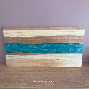 May include: A wooden serving board with a teal blue epoxy resin inlay. The resin has a marbled effect. The board is rectangular and has a natural wood finish.