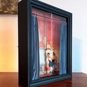 May include: A framed artwork depicting two figures performing a trapeze act in a theater setting. The figures are suspended from the ceiling by ropes, and the background features red and blue curtains.