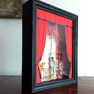 Puede incluir: Una caja de sombra enmarcada en negro con un tel&oacute;n de fondo de cortina roja y dos figuras actuando en un escenario. Las figuras son un hombre y una mujer, ambos vestidos con trajes coloridos. El escenario est&aacute; decorado con detalles ornamentados.