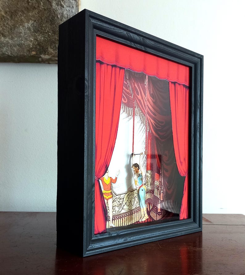 Puede incluir: Una caja de sombra enmarcada en negro con un tel&oacute;n de fondo de cortina roja y dos figuras actuando en un escenario. Las figuras son un hombre y una mujer, ambos vestidos con trajes coloridos. El escenario est&aacute; decorado con detalles ornamentados.