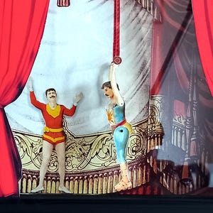 Puede incluir: Dos figuras de artistas de circo en un escenario. Una figura es un hombre con un traje rojo y amarillo con los brazos en alto. La otra figura es una mujer con un traje azul y blanco colgando de una cuerda roja. El escenario est&aacute; decorado con ribetes dorados y rojos.