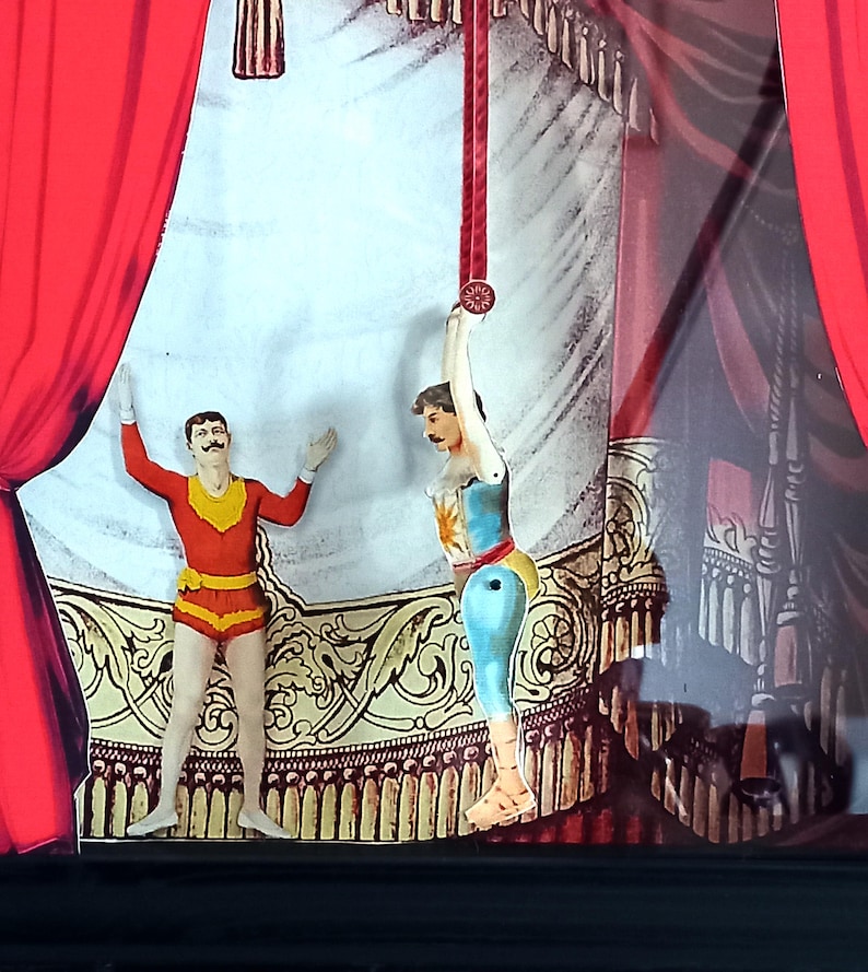 Puede incluir: Dos figuras de artistas de circo en un escenario. Una figura es un hombre con un traje rojo y amarillo con los brazos en alto. La otra figura es una mujer con un traje azul y blanco colgando de una cuerda roja. El escenario est&aacute; decorado con ribetes dorados y rojos.