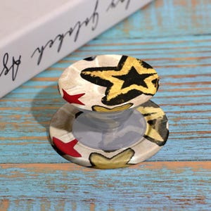 May include: A round phone grip with a star pattern in gold, black, and red on a cream background. The grip is attached to a clear, collapsible base. The background is a blue and brown wooden surface.