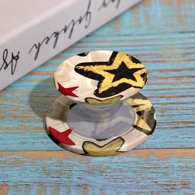 May include: A round phone grip with a star pattern in gold, black, and red on a cream background. The grip is attached to a clear, collapsible base. The background is a blue and brown wooden surface.