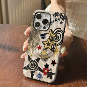 May include: A phone case with a beige marble pattern and a collage of star and spiral designs in black, gold, red, and blue. The case has a clear pop socket and a camera cutout.
